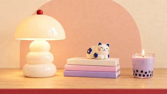 Cream lamp, pastel books, ceramic cat, and taro boba candle on an oak table with a peach arch backdrop